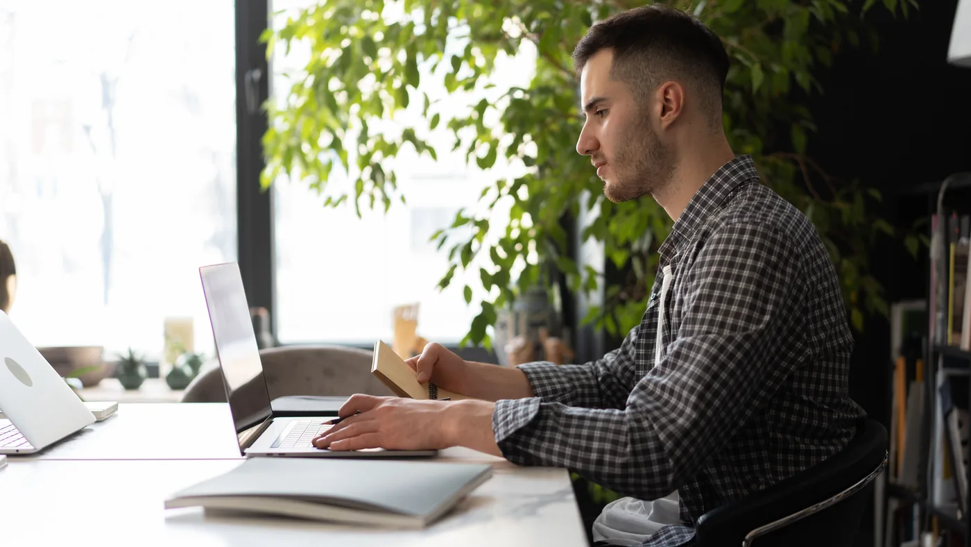 A man at a laptop desk in a bright, plant-filled office, notebook in hand